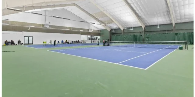 Indoor tennis court featuring blue playing surface, surrounded by spectators and training areas, relevant to racquet sports and wellness activities at The DRHC.